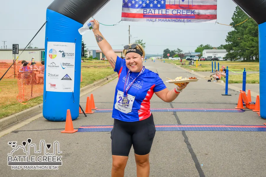 A smiling woman in a blue and red race shirt crosses the finish line of the Battle Creek Half Marathon & 5K, holding a plate of food and a bottle of water.