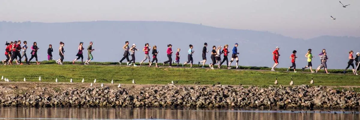 A group of runners race along a path next to a body of water, with hills in the background.
