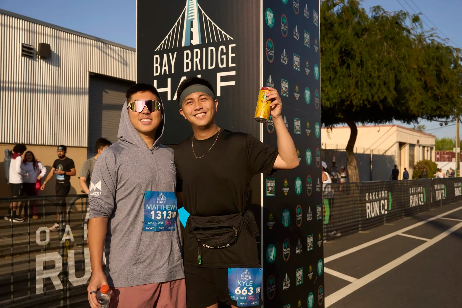 Two smiling men pose for a photo in front of a Bay Bridge Half Marathon sign, one holding a can of Yerba Mate.