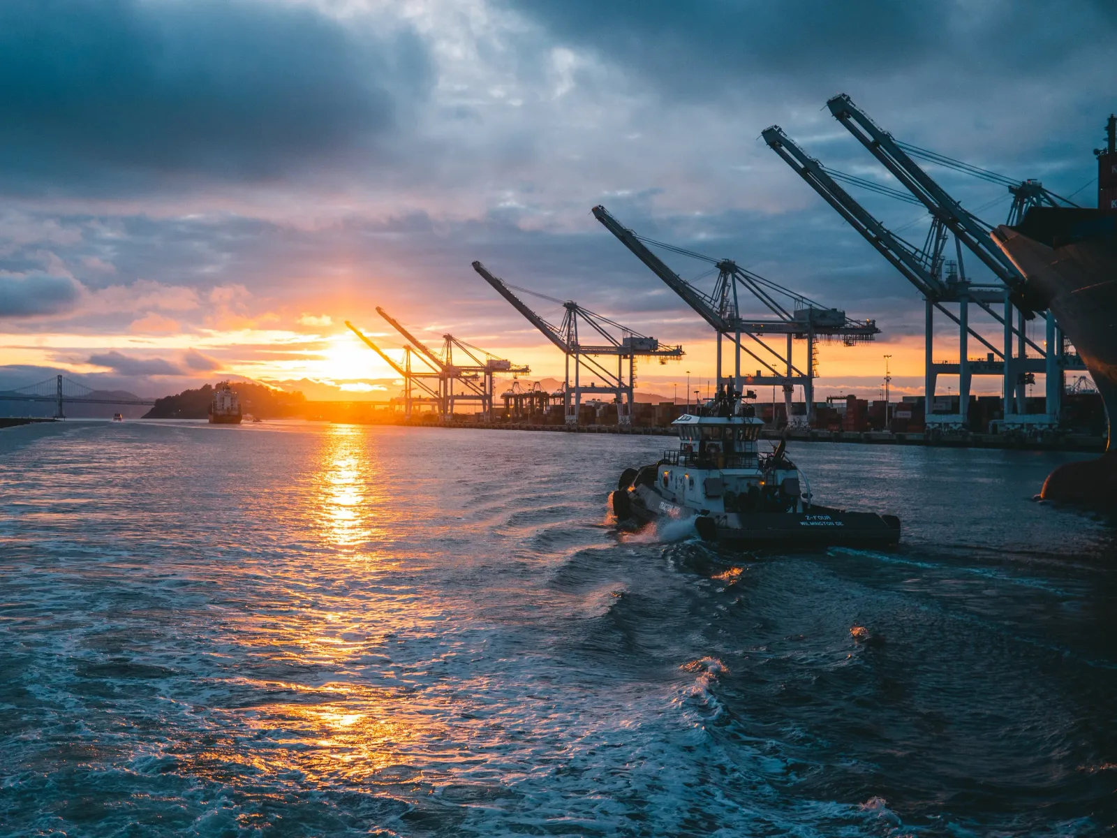 A tugboat navigates through choppy water with a bridge and industrial cranes in the background during a dramatic sunset.