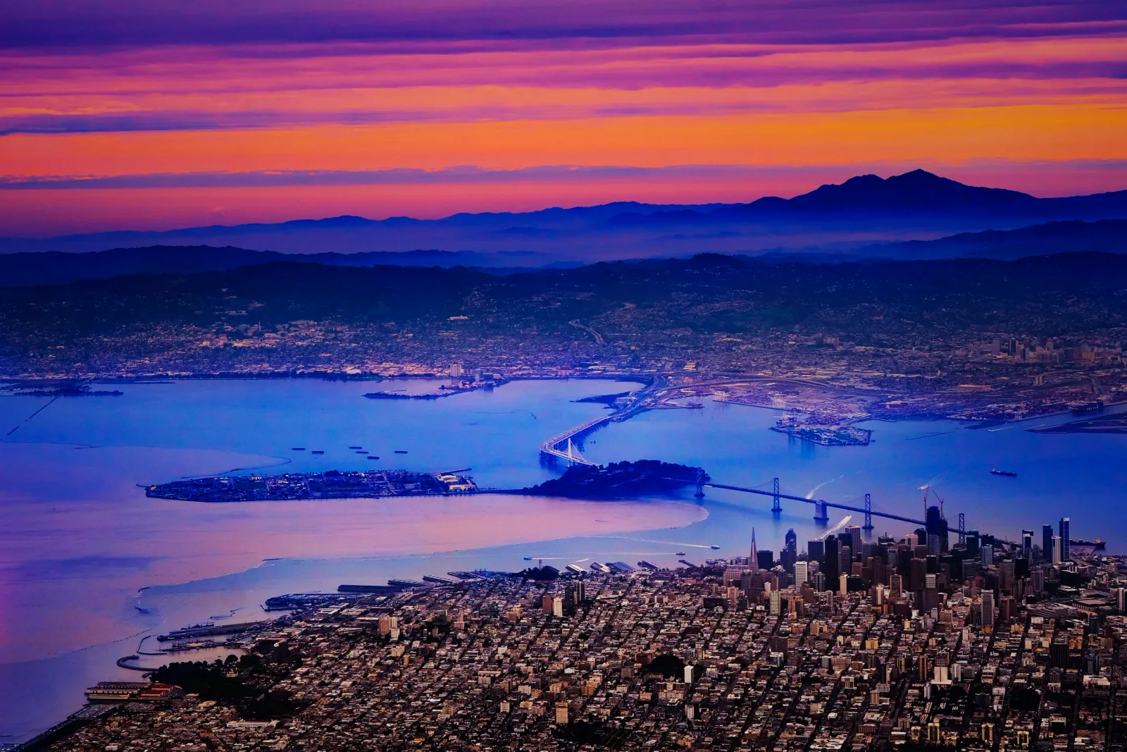 Aerial view of the San Francisco Bay and Bay Bridge at sunset, with the city skyline in the foreground and mountains in the background.