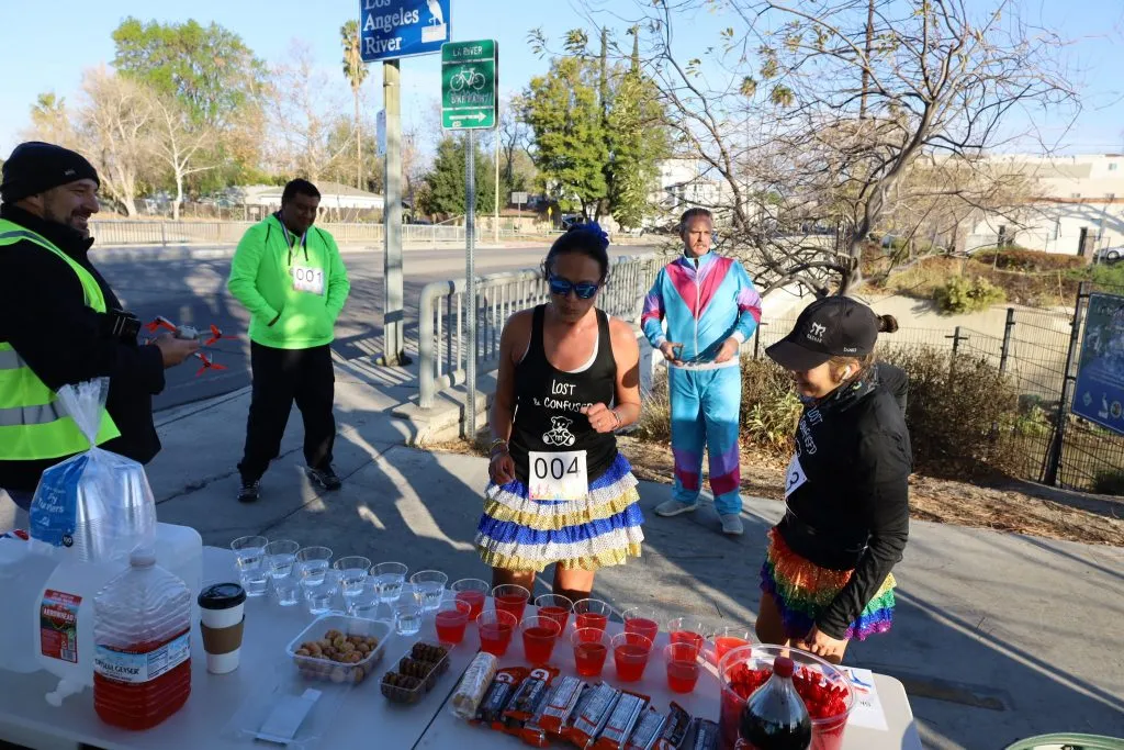 Runners and volunteers at an aid station with drinks and snacks, with signs for the Los Angeles River in the background.