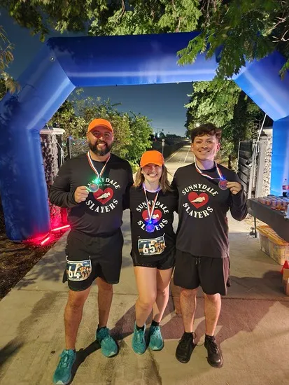 Three smiling runners wearing matching shirts and holding medals stand under a blue inflatable archway.