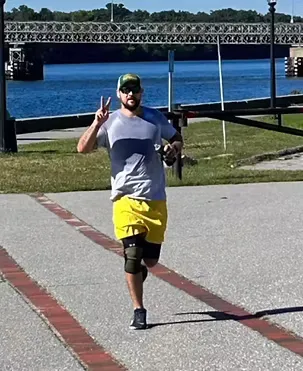 A runner wearing a grey t-shirt, yellow shorts, and a hat runs on a paved path next to a body of water, holding up two fingers in a peace sign.