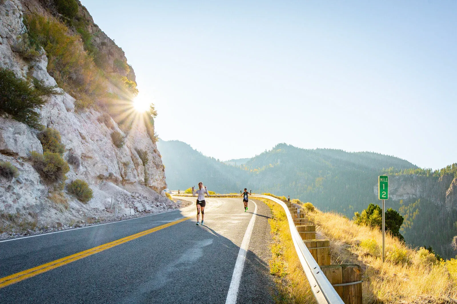 Two runners on a winding road with a mountain backdrop and sun flare.