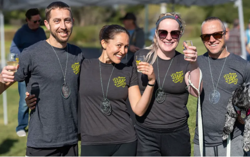 A group of four people, two men and two women, smiling and holding drinks, wearing dark t-shirts with a 'Festival Beer City' logo and clear oval medals.