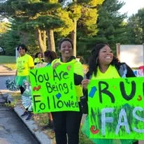 Two women in neon shirts smile and hold large signs, one reading "You Are Being Followed" and the other partially visible with "RUN FASTER".