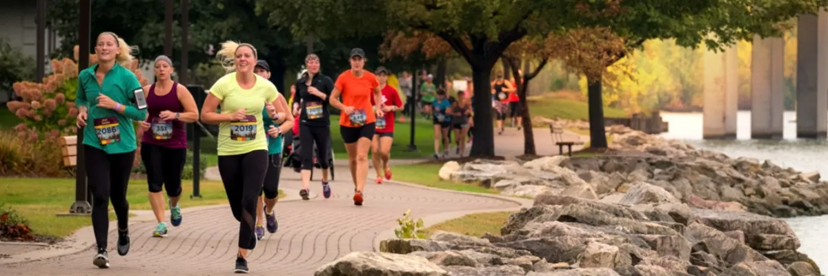 A group of women running along a paved path next to a body of water, with trees and a bridge in the background.
