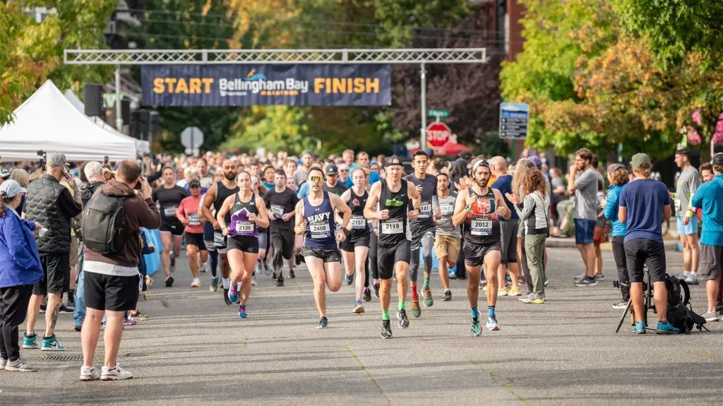 Runners starting the Bellingham Bay Marathon under a banner that reads 'START Bellingham Bay FINISH'.