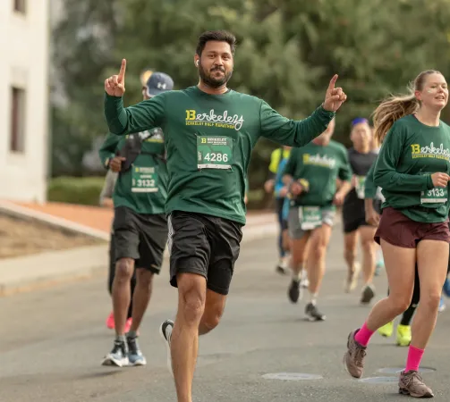 A male runner in a green Berkeley Half Marathon shirt celebrates with his arms raised while running in a race.