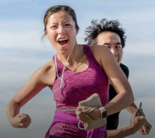 A female runner with an excited expression, with a male runner behind her, both appearing to be in motion.