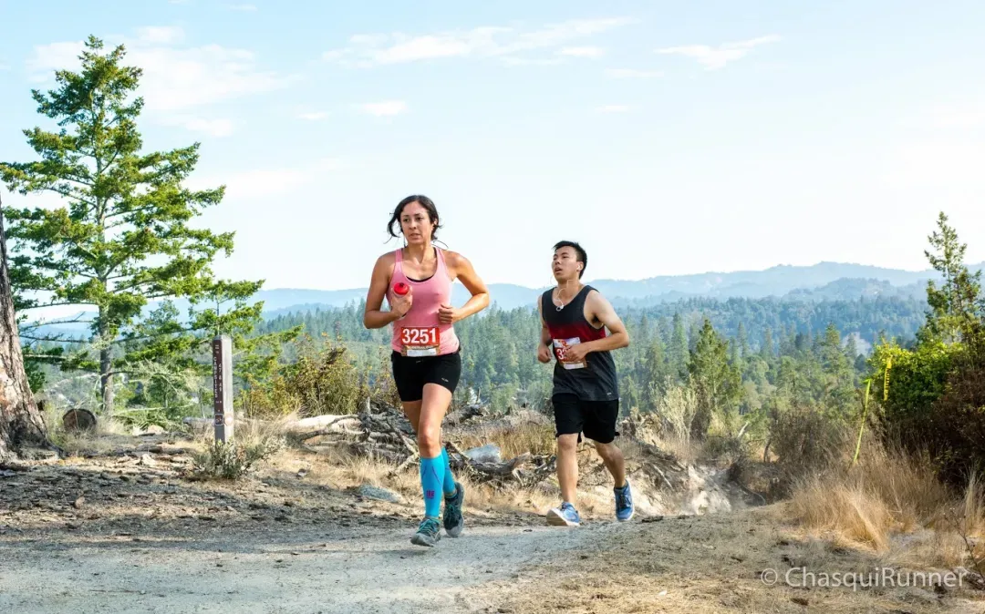 Two runners, a woman in the foreground and a man behind her, running on a dirt trail with a scenic view of rolling hills and trees in the background.