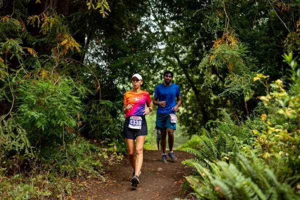 Two runners on a dirt trail surrounded by lush green forest.