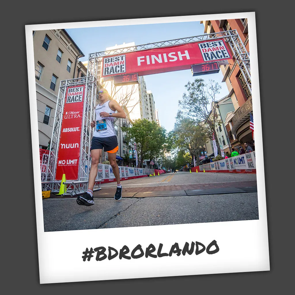 A runner crosses the finish line at the Best Damn Race Orlando, with the finish banner overhead and city buildings in the background.