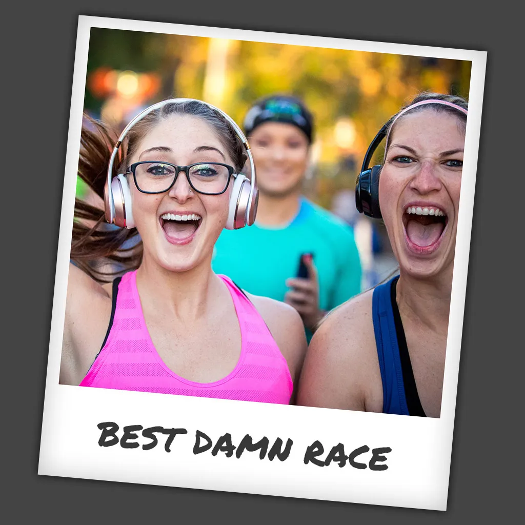 Two women with headphones on, smiling and shouting excitedly at a race, with another person blurred in the background.