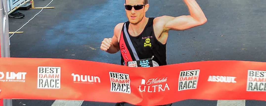 A male runner in sunglasses and a black tank top with a skull logo crosses the finish line, raising his arm in victory.