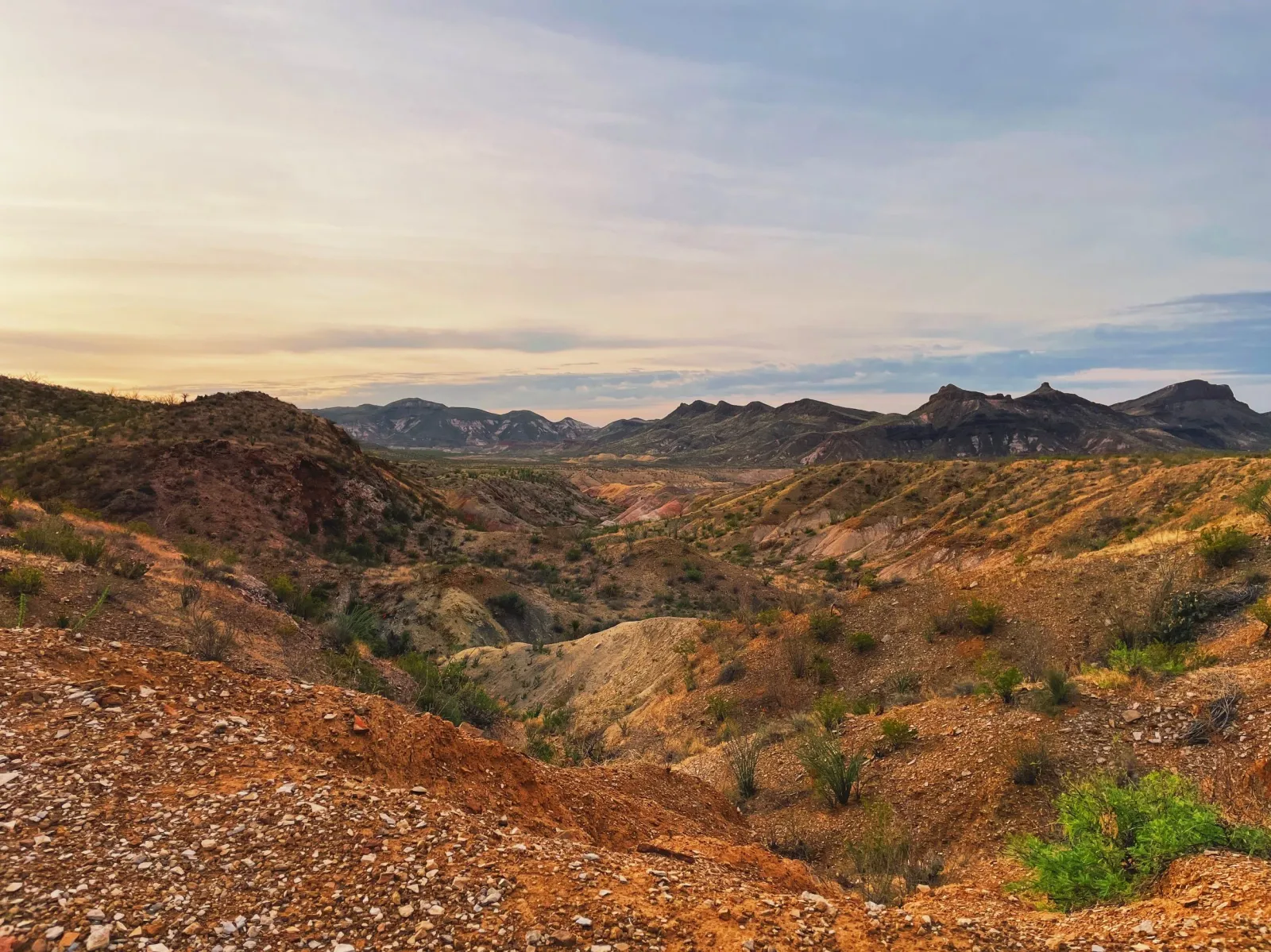 A wide scenic view of rolling desert hills and mountains under a cloudy sky at sunrise or sunset.