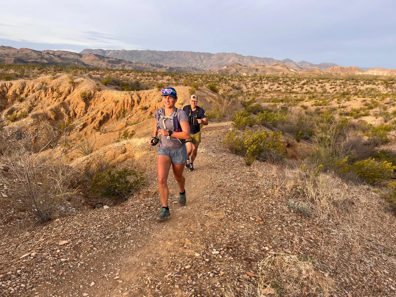 Two runners on a dusty trail with a mountainous desert landscape in the background.
