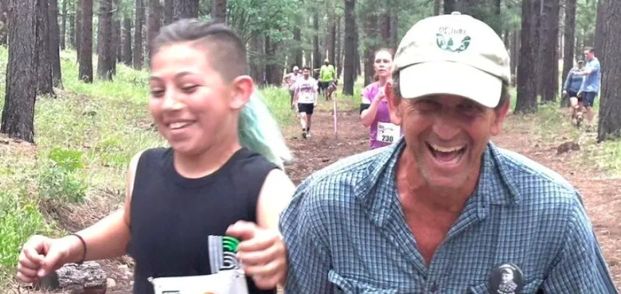 A young boy and an adult man laughing while running on a forest trail during a race.