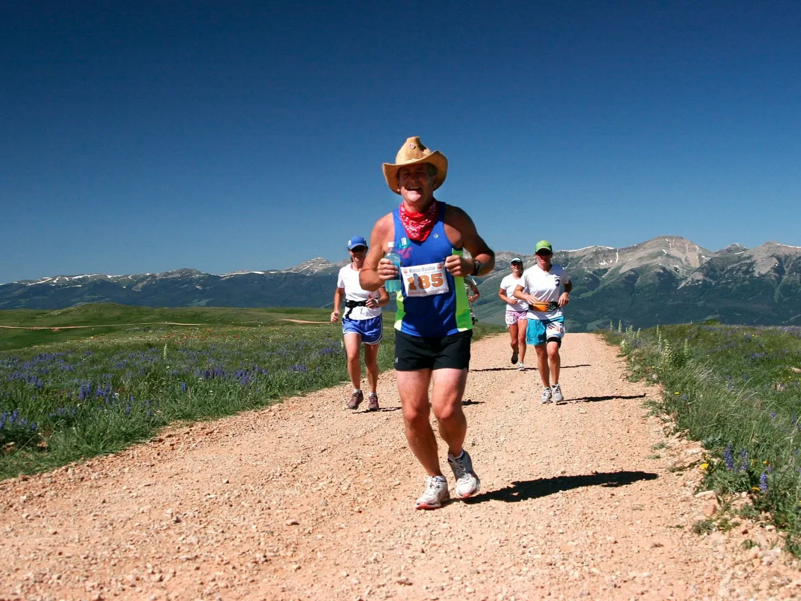 A runner wearing a cowboy hat and a red bandana runs on a dirt road with mountains in the background, followed by other runners.