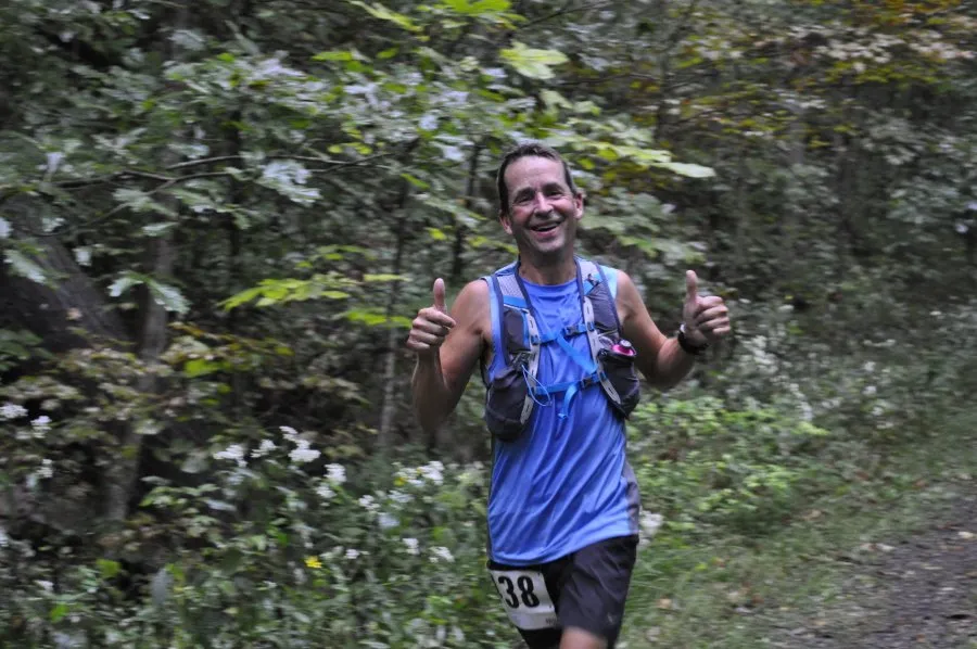 A male runner with bib number 38 smiles and gives two thumbs up while running on a wooded trail.