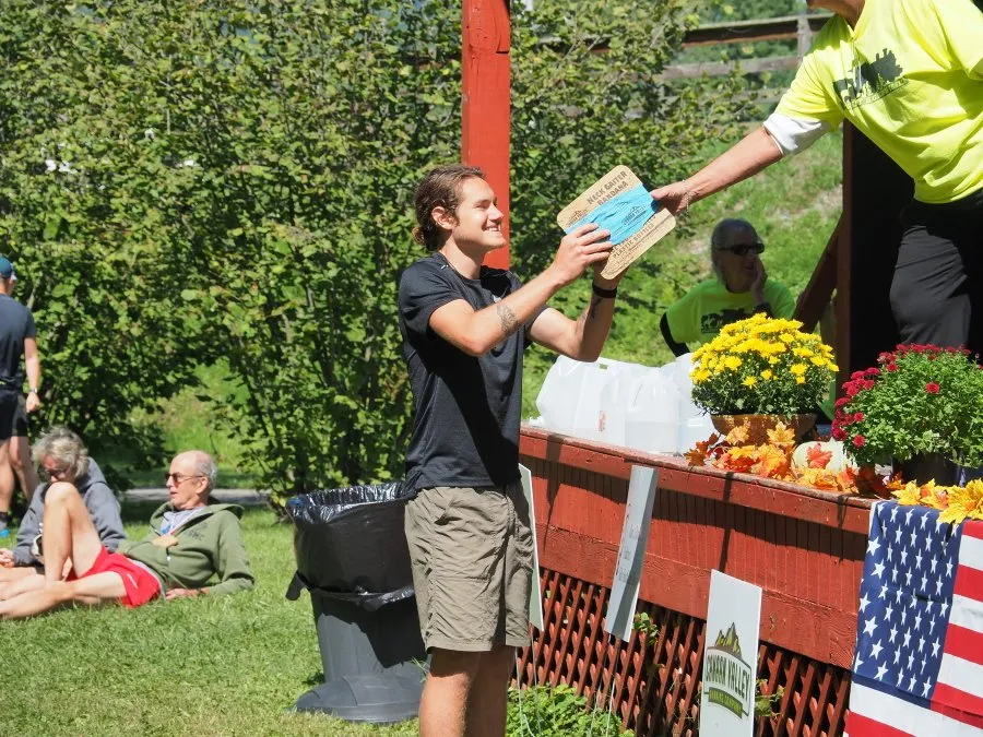A runner receiving a neck gaiter from a volunteer at a race aid station, with other participants relaxing in the background.