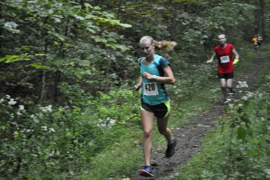 A female runner with bib number 420 runs on a wooded trail, with another runner visible in the background.