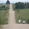 Two runners ascend a long, dusty hill on a dirt road, with green fields and trees on either side.