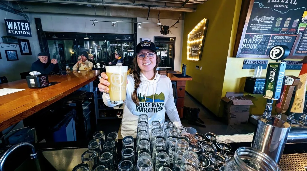 A smiling woman wearing a Boise River Marathon hoodie holds up a glass of beer.