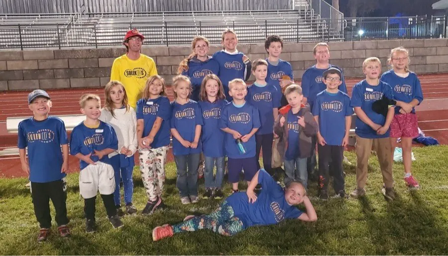 A group of children and adults in blue t-shirts with a 'Solekids' logo, posing for a photo on a grassy field in front of stadium seating.