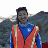 A woman smiles at the camera, wearing a blue race jacket and an orange safety vest, with a desert landscape in the background.