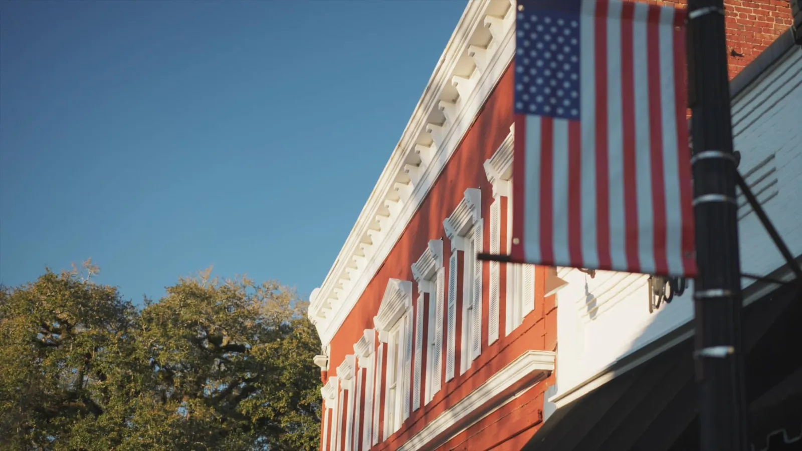 A red building with white trim and windows, an American flag hanging from a pole, and trees against a clear blue sky.