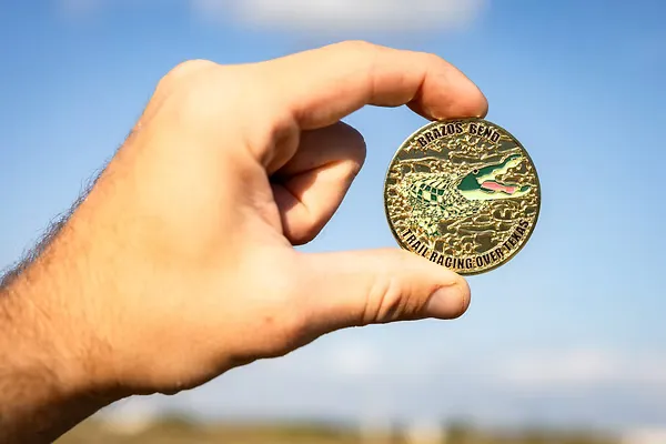 A hand holds up a gold medal with an alligator design and the text 'BRAZOS BEND TRAIL RACING OVER TEXAS' against a blue sky.