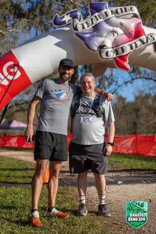 Two male runners smiling and posing for a photo under the Brazos Bend 100 Trail Racing Over Texas arch.