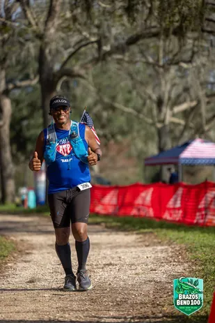 A male runner smiles and gives two thumbs up while running on a dirt path, with an American flag attached to his backpack.