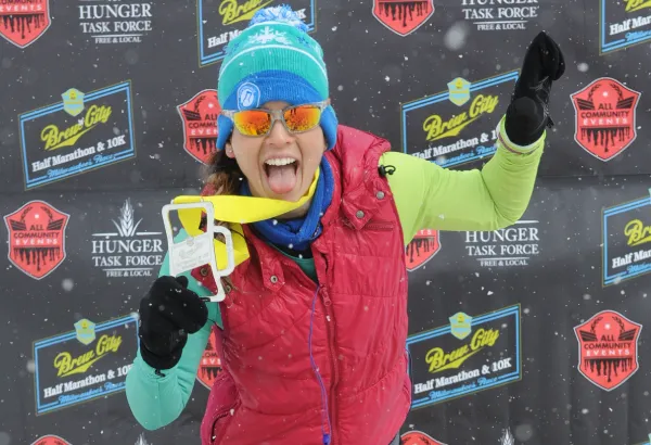 A runner celebrates with a medal and a playful expression in front of a backdrop with race branding.