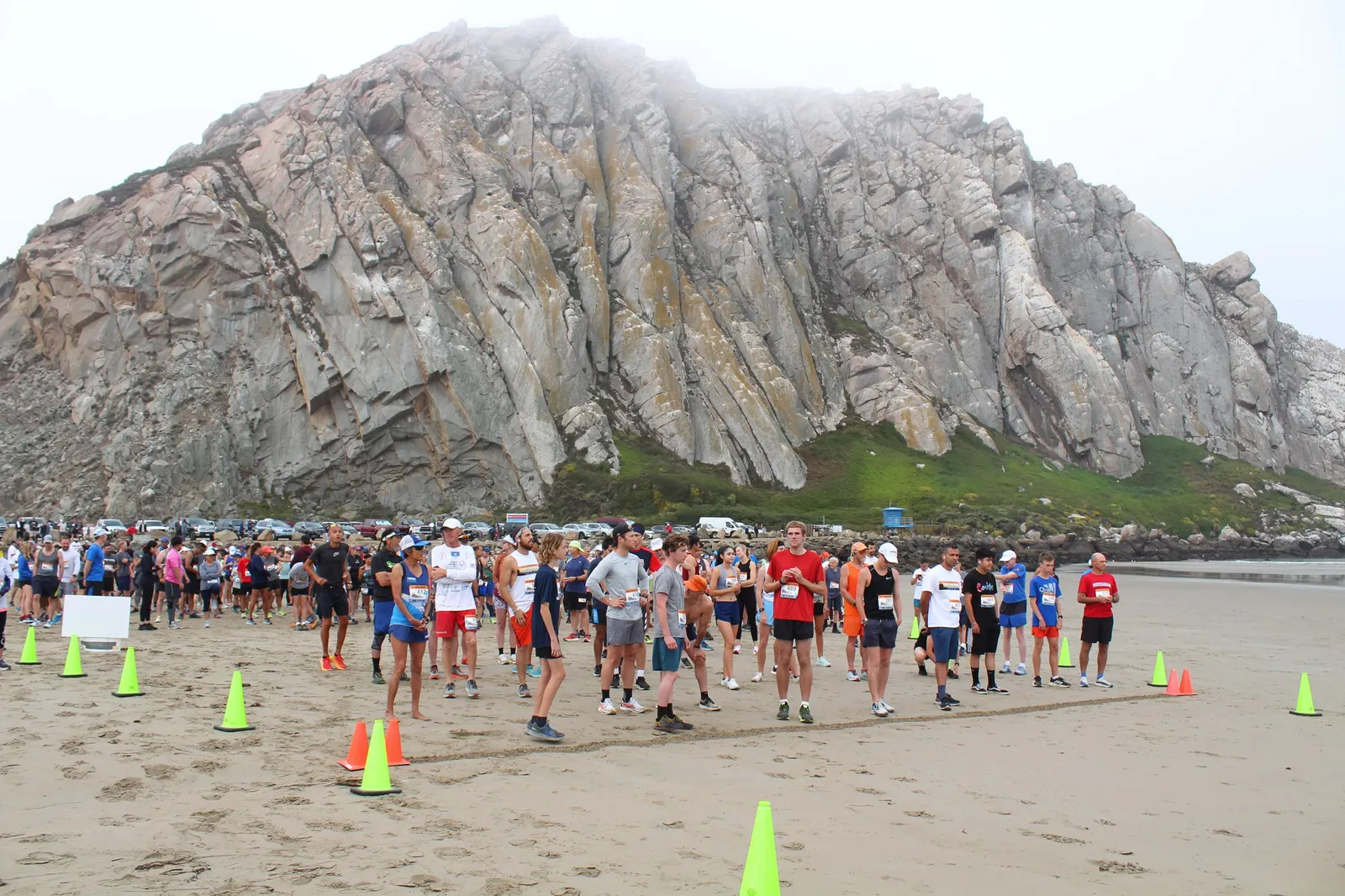 A crowd of runners stand on a sandy beach, with a large, imposing rock formation behind them and cars parked in the distance. The runners are facing forward, appearing to be at the start line of a race.