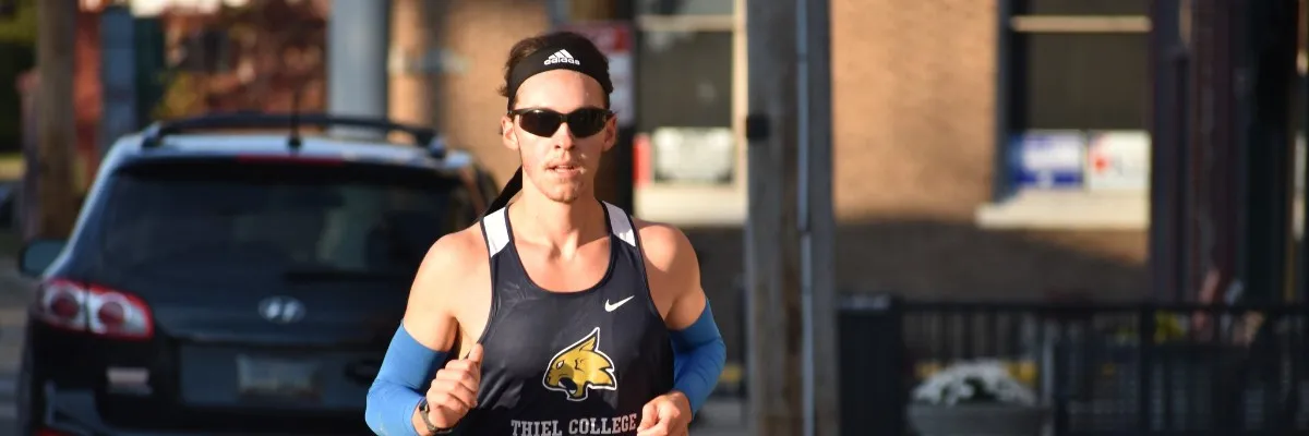 A runner wearing sunglasses and a headband, with a 'Thiel College' singlet, running on a road with a car in the background.