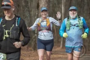 Three runners on a wooded trail, with the middle runner waving at the camera.