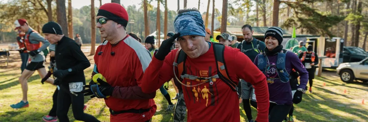 Runners in athletic wear, some wearing hats and gloves, are starting a race in a wooded area. One runner in the foreground salutes.