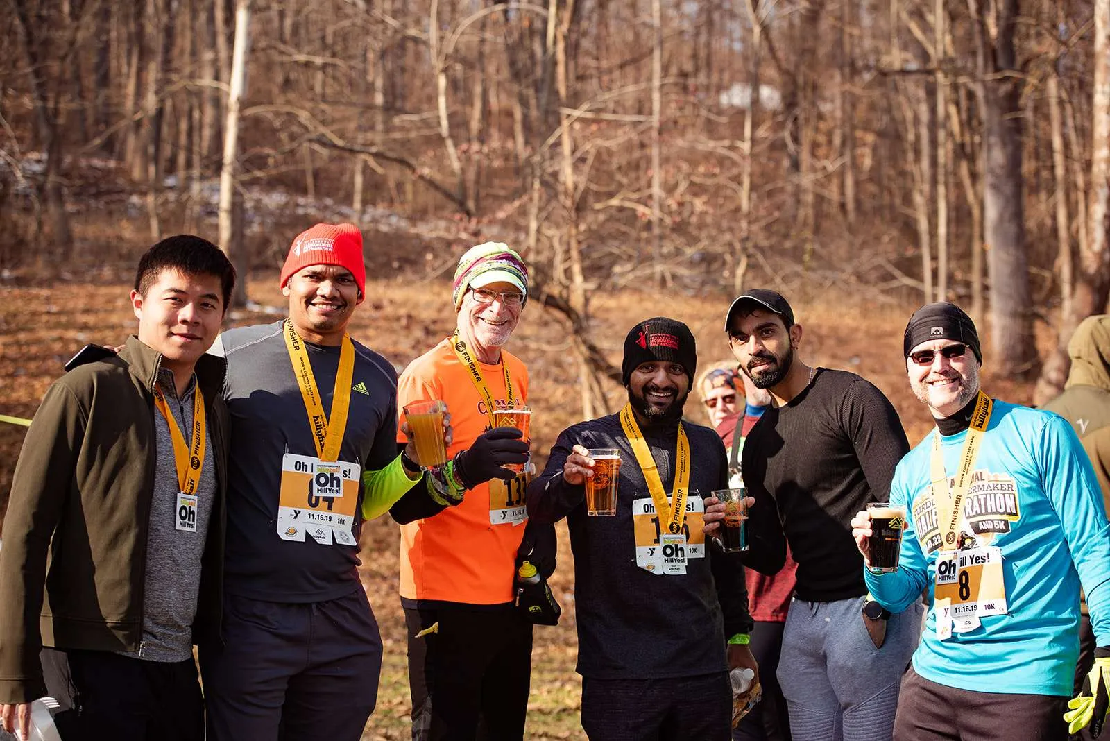 A group of six men, all wearing finisher medals and smiling, some holding drinks, in a wooded outdoor setting after a race.