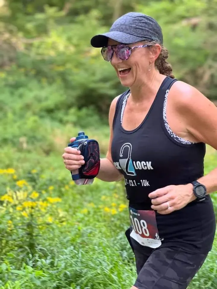 A smiling female runner wearing a black tank top with a race logo and bib number 108, running on a trail with yellow flowers in the background.