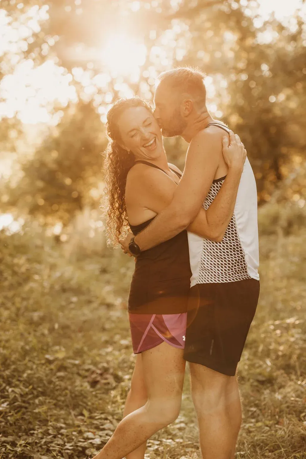 A man kisses a woman on the cheek as they embrace, both wearing running attire, with a golden sun flare in the background.