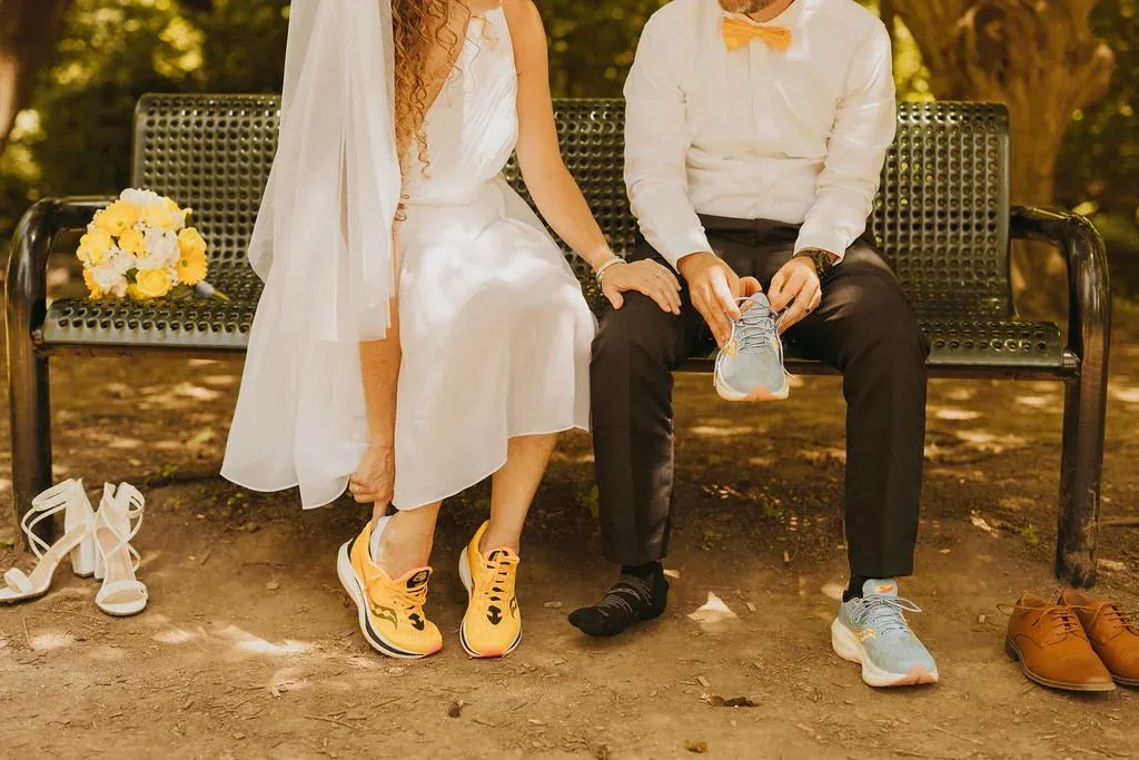A bride and groom in wedding attire sit on a park bench, both wearing running shoes, with a bouquet of yellow flowers beside them.