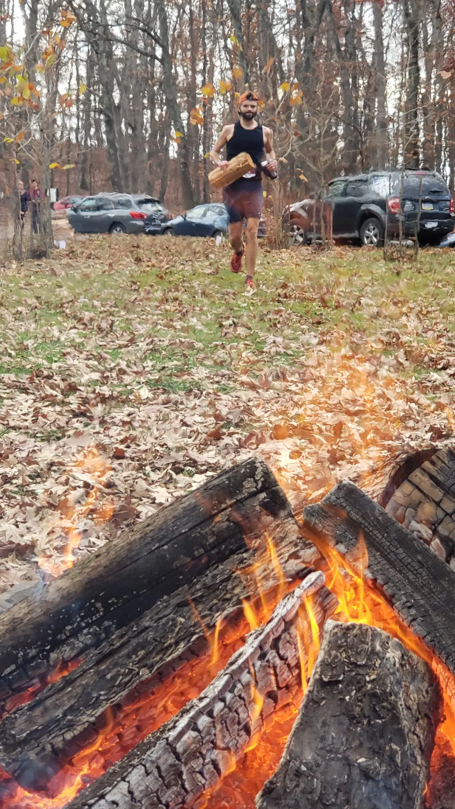 A male runner with a beard, wearing a black tank top and orange hat, runs through a forest with fallen leaves on the ground. A campfire burns in the foreground.