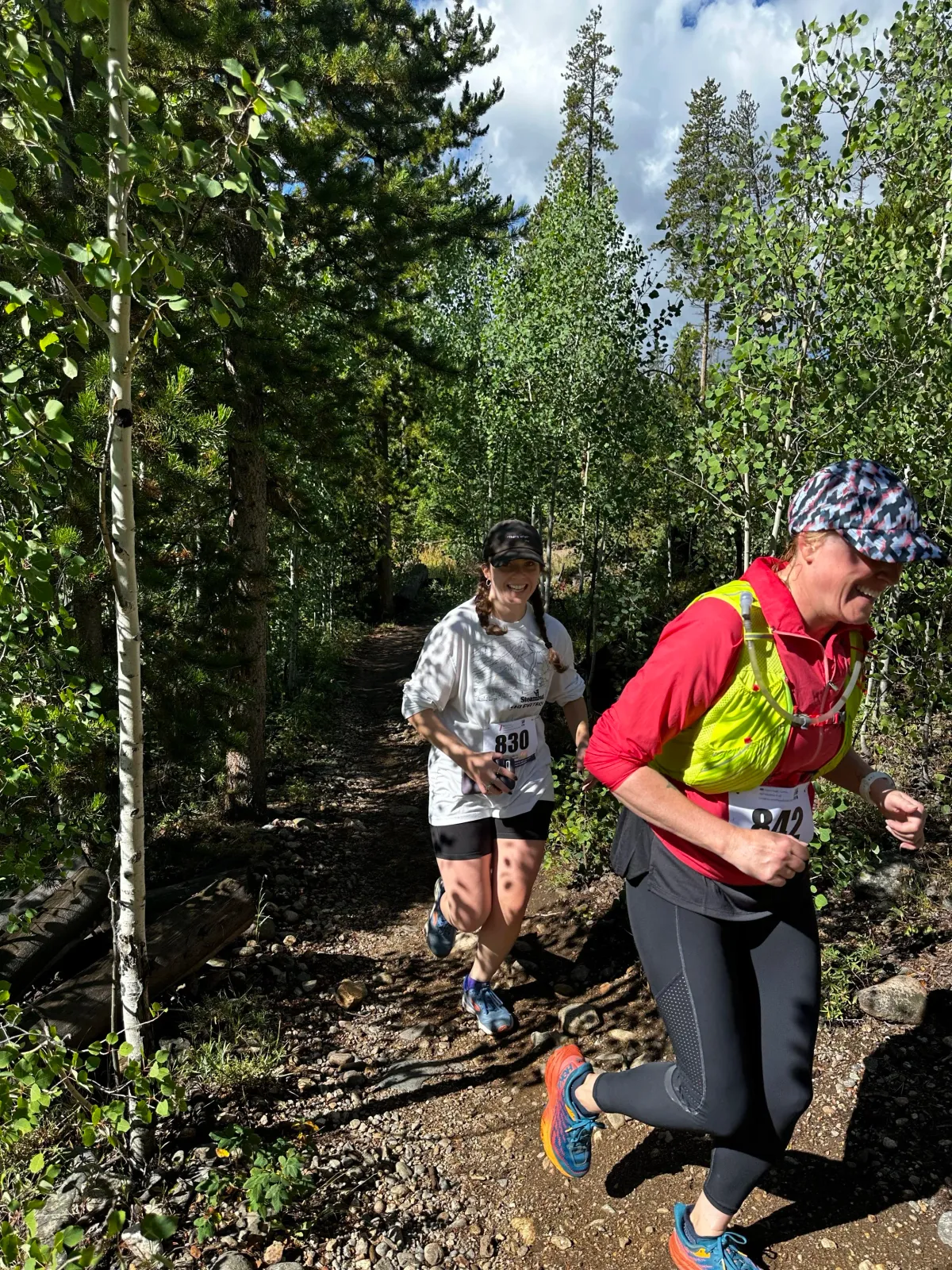Two runners on a wooded trail, one in the foreground wearing a red jacket and yellow vest, and another in the background wearing a white t-shirt.