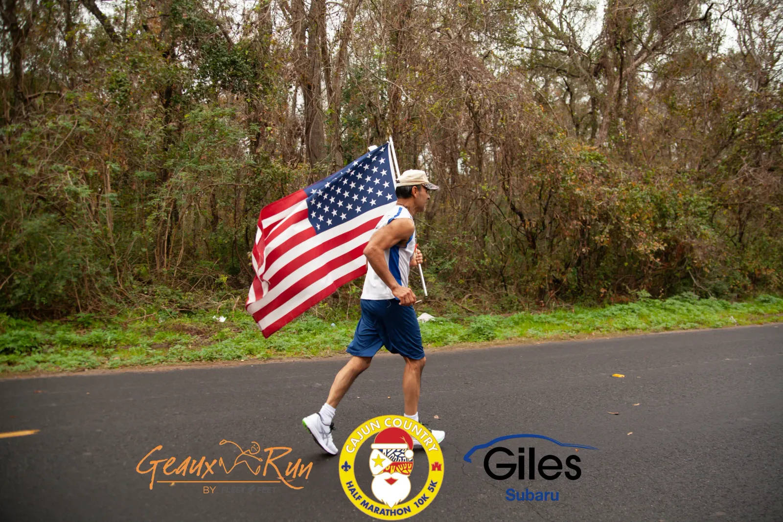 A male runner carries a large American flag while running on a road lined with trees.