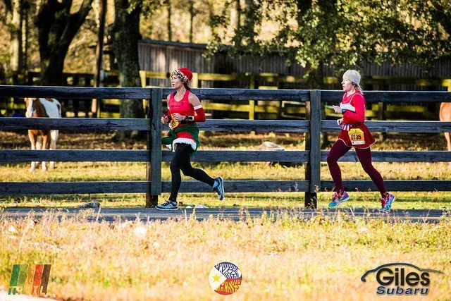 Two female runners in festive attire running on a road next to a wooden fence.
