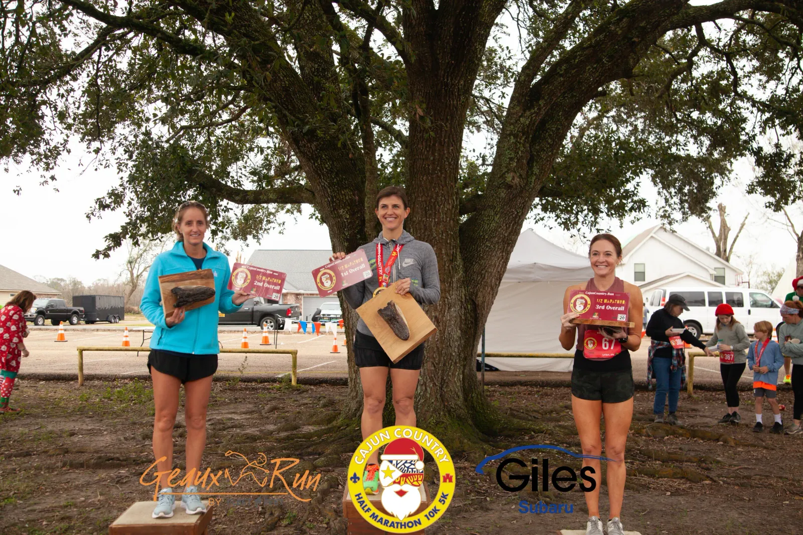 Three female runners on a podium holding awards and trophies, with a large tree in the background.