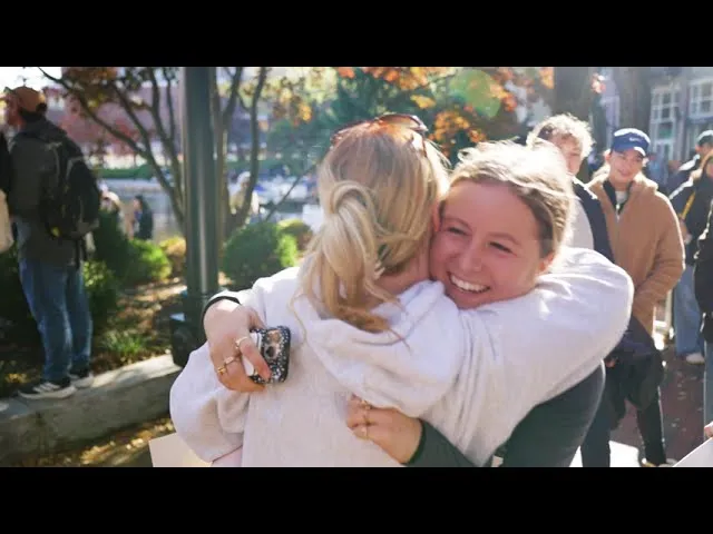 Two women hugging and smiling, with other people in the background at an outdoor event.